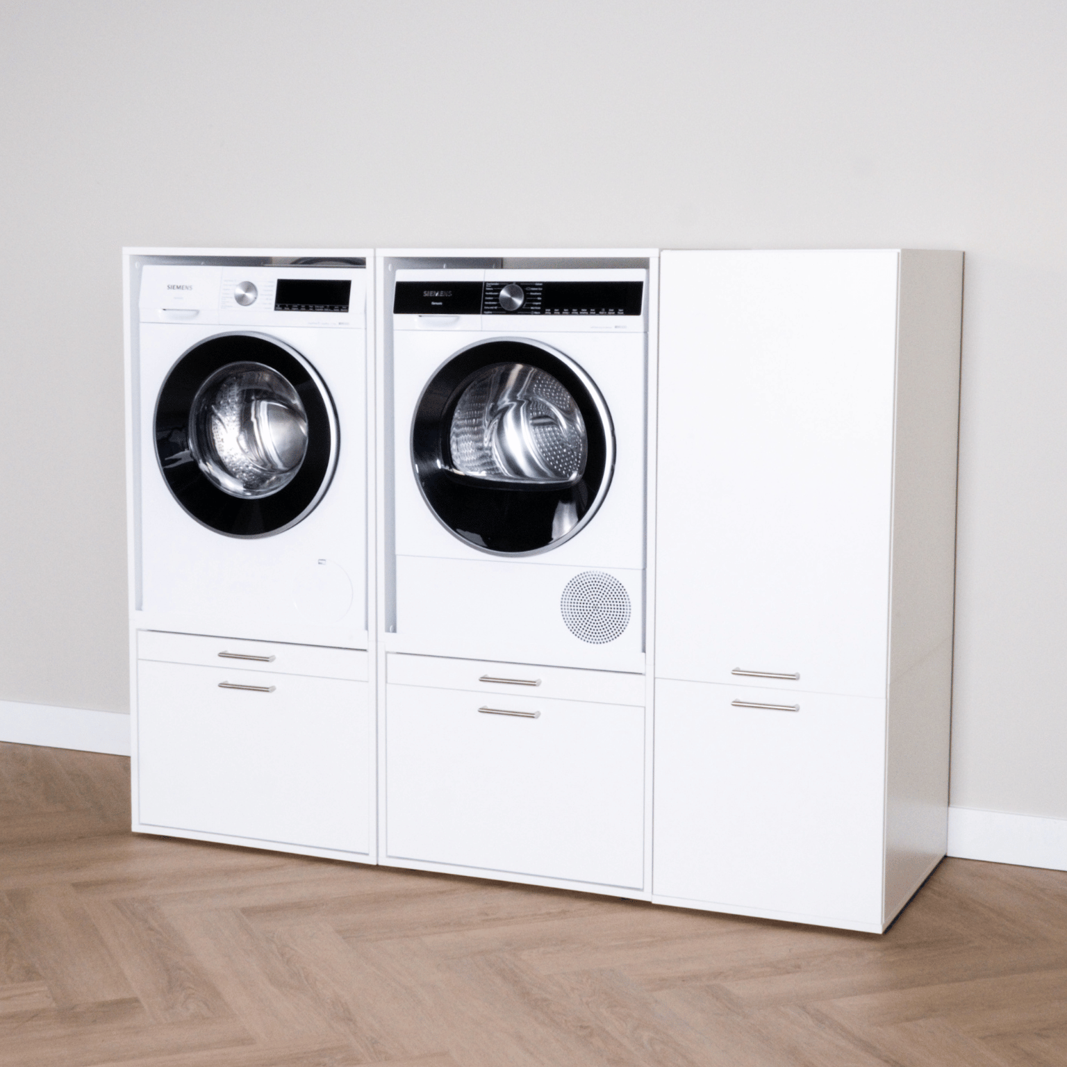 White front-load washer and dryer set with matching base drawers and tall storage cabinet on wood floor against gray wall.