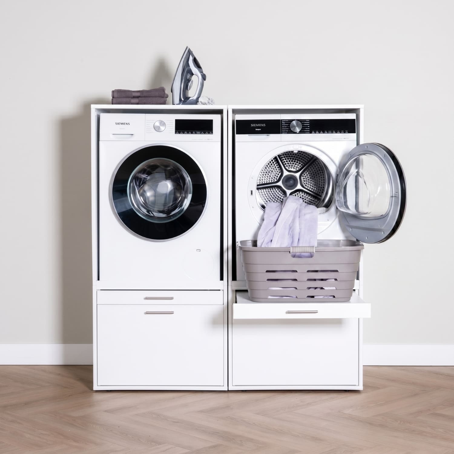 White front-load washer and dryer set with laundry basket on open dryer door and iron and folded clothes on top.