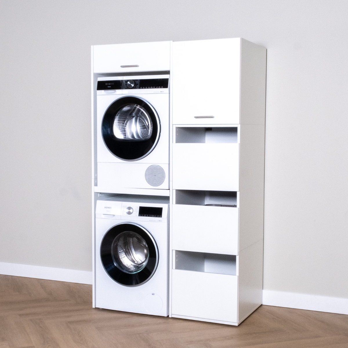 White stacked washer and dryer unit with tall white storage cabinet featuring open and closed compartments on hardwood floor.